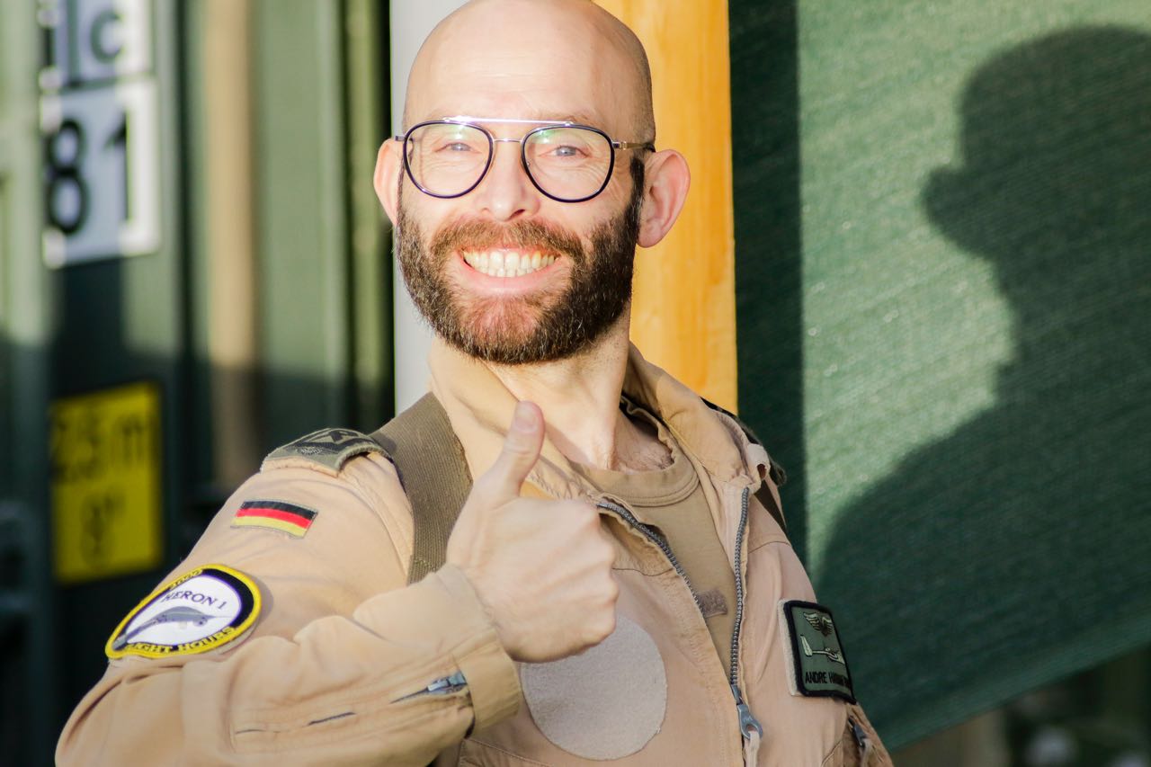 André Hassan Khan in Fliegerkombination, lächelnd mit erhobenem Daumen